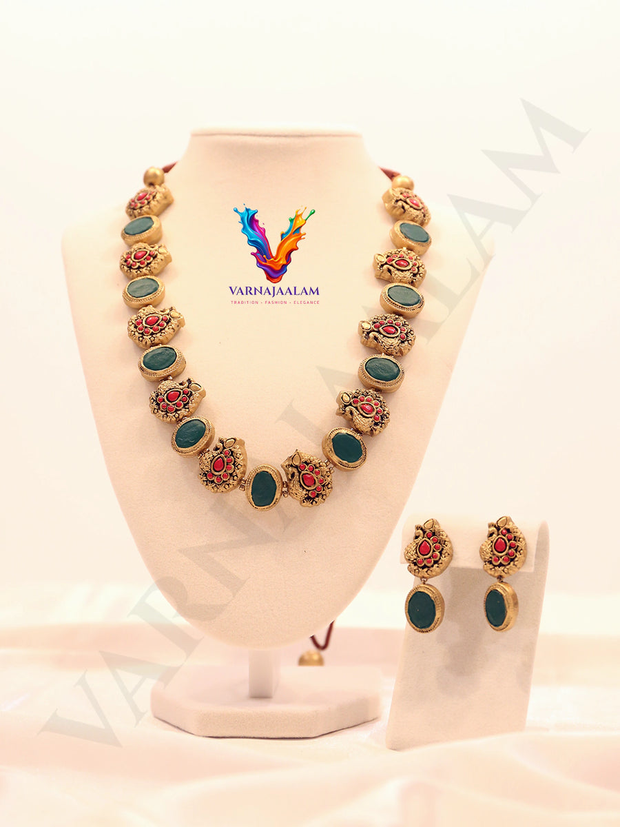 Handcrafted terracotta mala necklace set featuring alternating oval dark green stones and antique gold peacock motifs with red accents, with matching peacock drop earrings